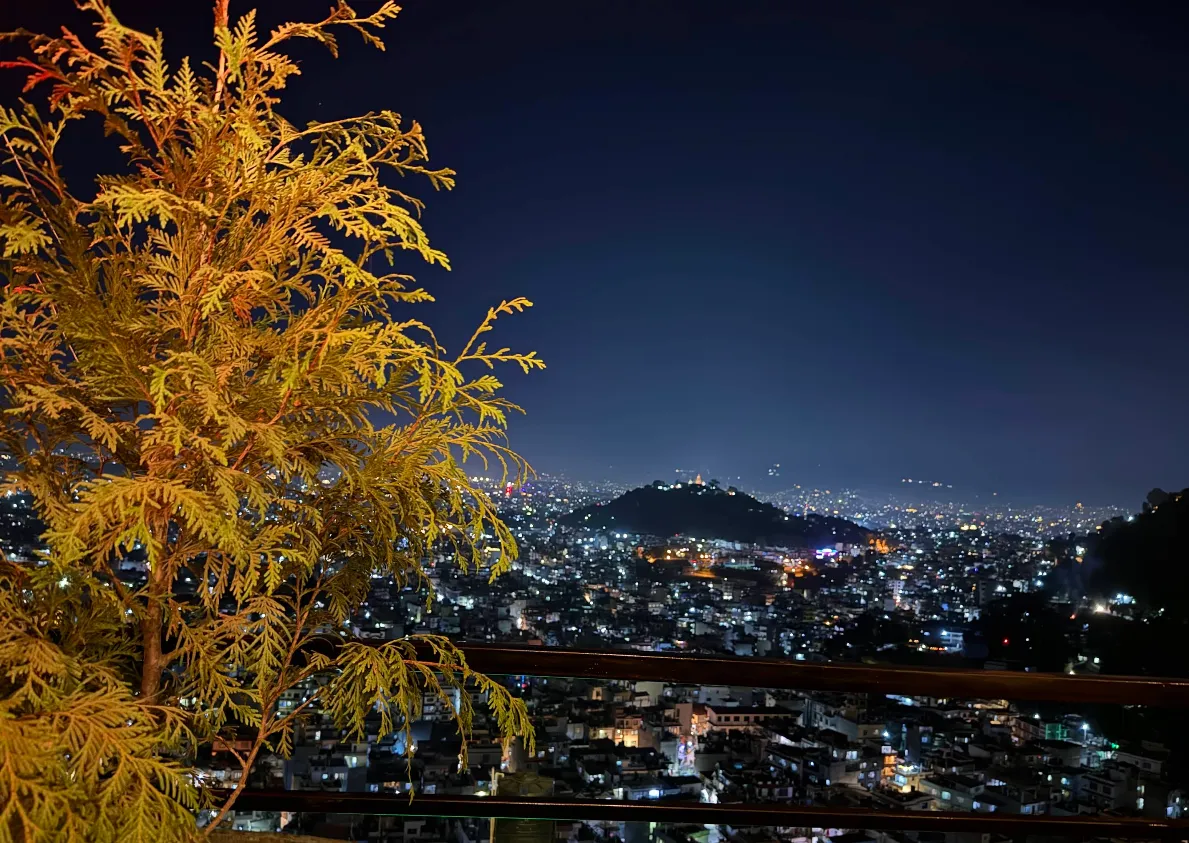 City lights of Kathmandu glowing beyond an amber-lit tree at the edge of the Skyline terrace
